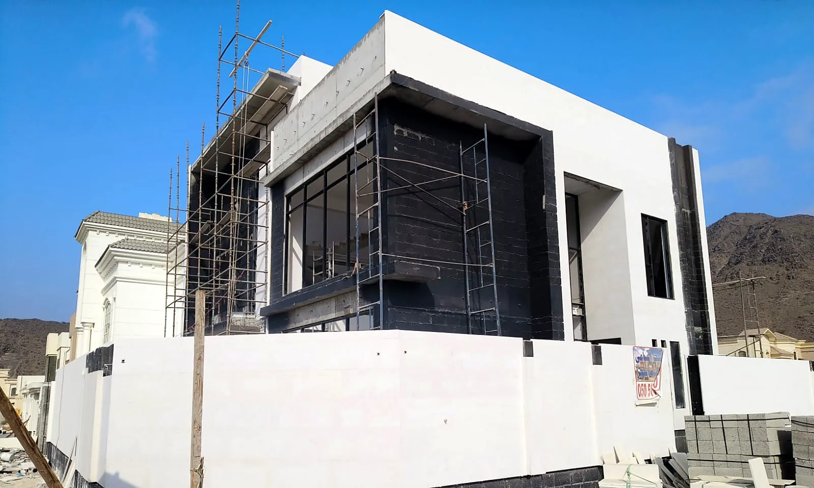 Corner view of a contemporary villa with black-framed glazing and scaffolding during façade works, mountains behind