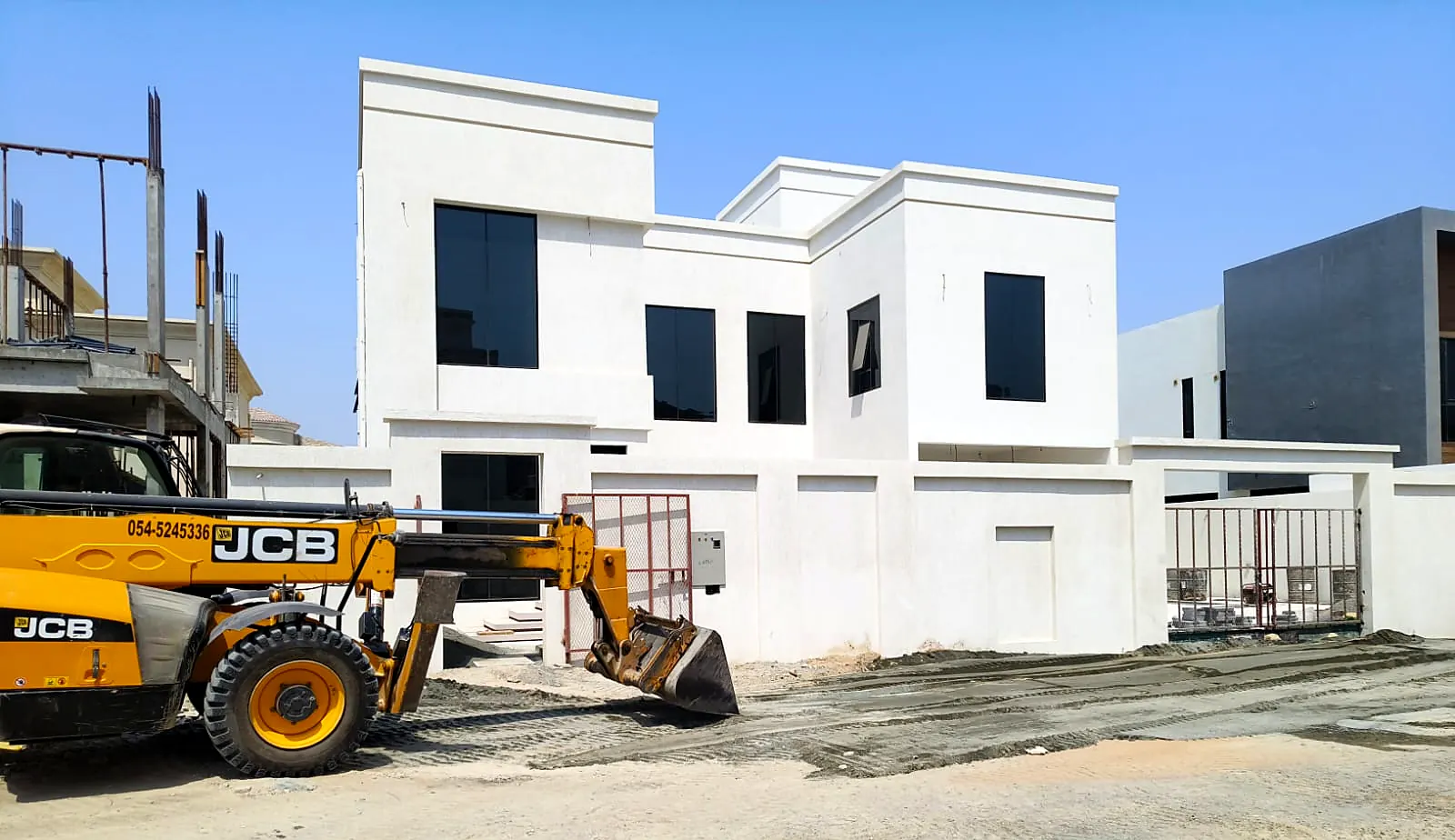 White two-storey villa with boundary wall while a loader carries out site works
