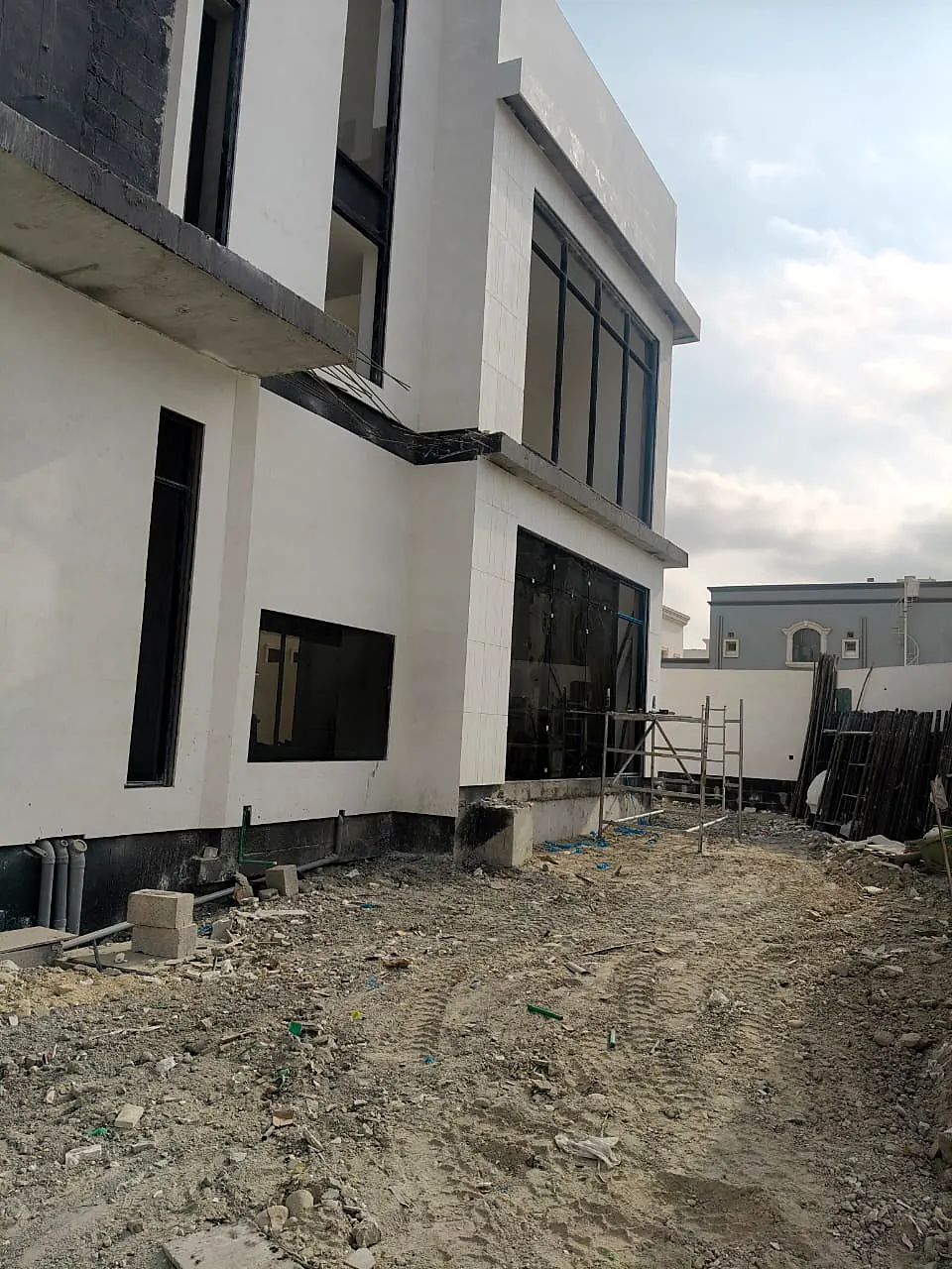 Side elevation of a white villa with dark stone bands and full-height windows while site levelling continues
